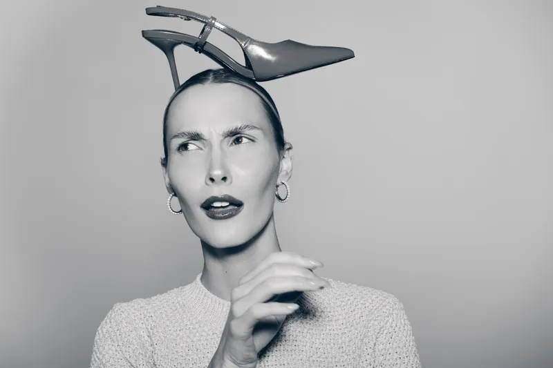 Confused white woman with pale skin and slicked hair holds high heel shoe on head, wearing earrings and knit sweater in monochrome studio portrait.