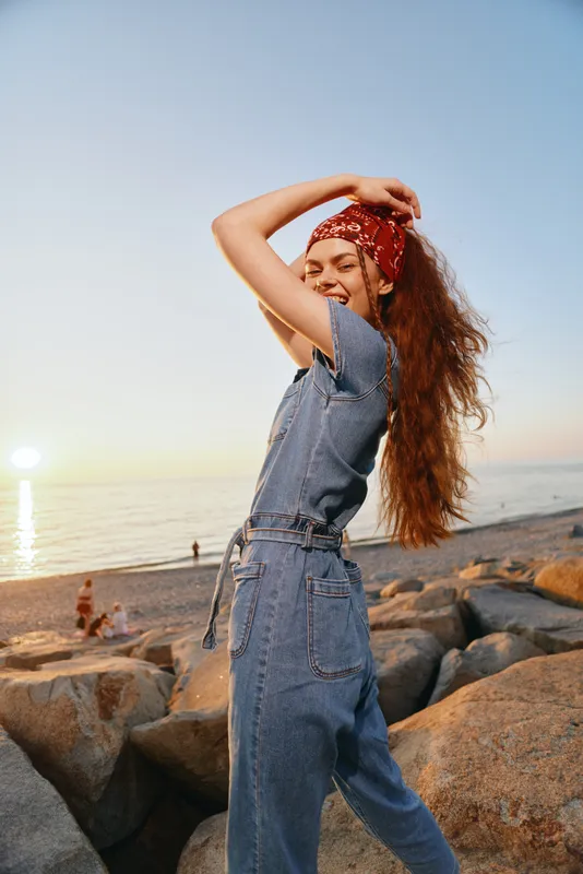 smiling woman in denim jumpsuit with red bandana poses joyfully at rocky beach during golden hour sunset warm natural light