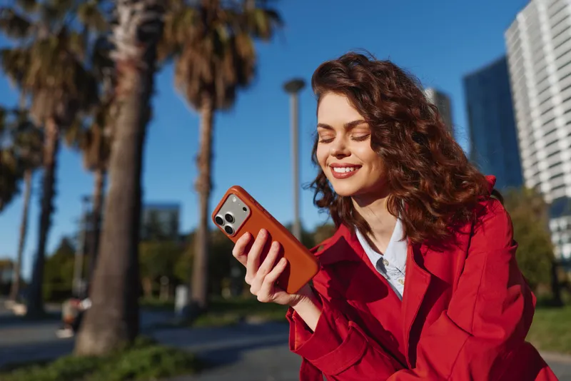 Young woman with curly hair smiles as she looks at her smartphone while sitting outdoors in a red jacket on a sunny day.