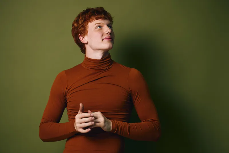 Young man with red hair and fair skin wearing burnt orange turtleneck looks away thoughtfully against green background. Casual style, studio portrait with soft lighting and shadow.