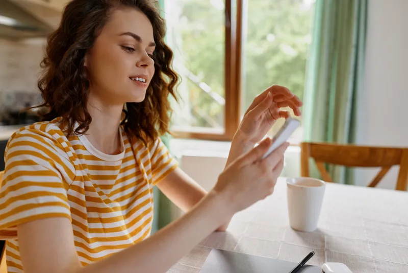 Young woman with wavy brunette hair sits at a bright home table, scrolling on a smartphone with a gentle smile, conveying a relaxed everyday lifestyle moment in a warm, cozy kitchen with natural