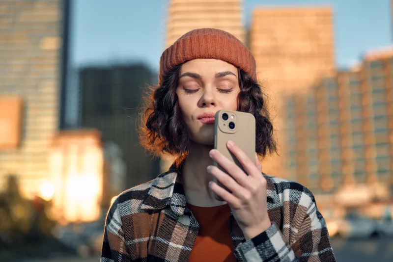 Woman with smartphone in urban setting during goldenhour, beanie and plaid jacket, candid portrait capturing mindfulness and authenticity with emotional storytelling and warm glow.