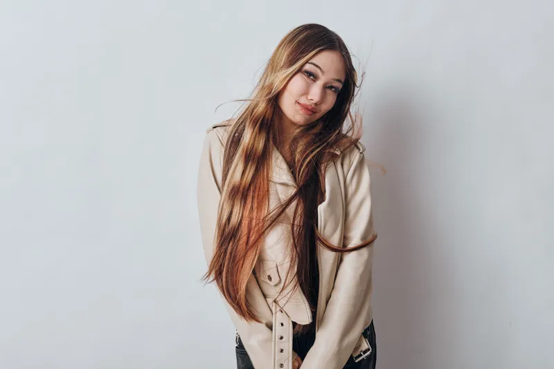 Young woman with long hair wearing beige jacket stands against white background looking at camera with gentle expression, fashion style and portrait concept.