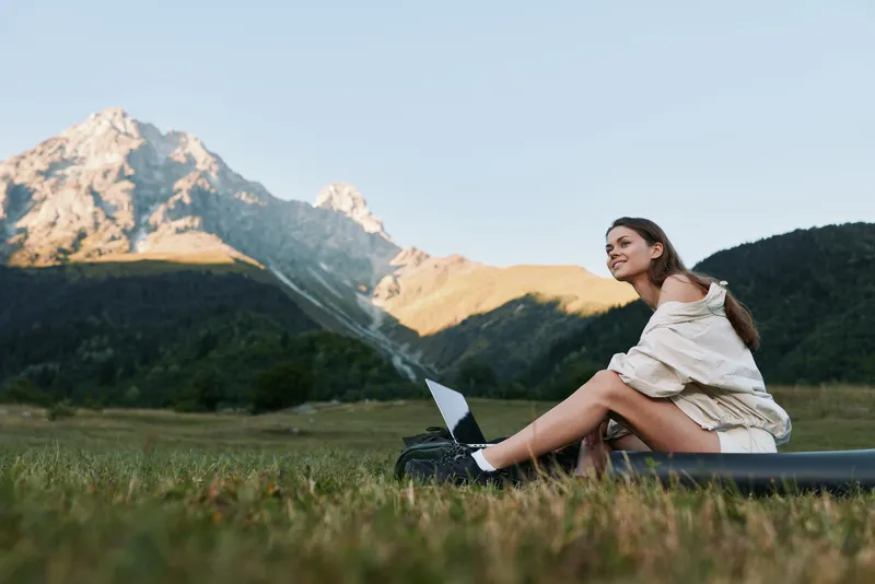 A cheerful young woman sits on a grassy field with a laptop, surrounded by mountains at sunset. She smiles softly, focused on her screen, exuding calm concentration and creative energy in a peaceful