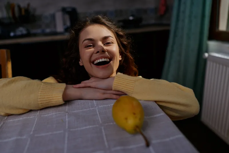 Cheerful young woman indoors rests her chin on folded arms across a dining table, smiling at a pear in the foreground as warm light creates a cozy scene.