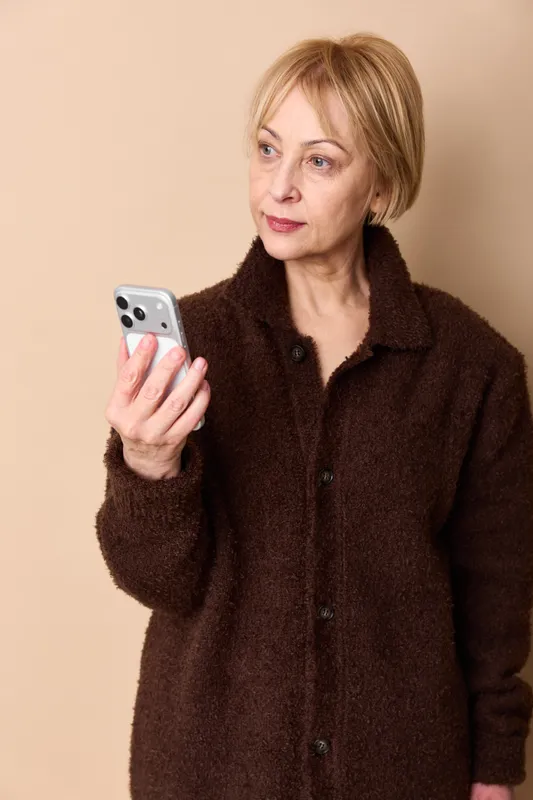 Elderly woman holds smartphone for communication in an indoor portrait, neutral expression, warm light against beige background, showing technology use and calm.