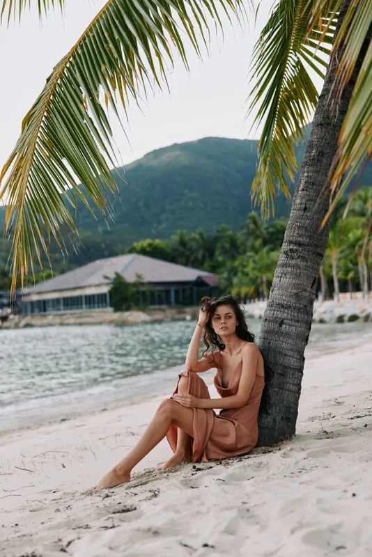 Relaxed woman in beige dress sitting on the sandy beach near palm tree with tropical mountain and water background under clear sky.