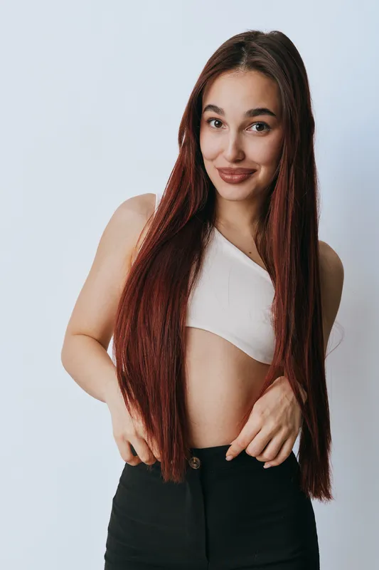 Young woman with long straight hair wearing white crop top and black pants posing against light background with confident smile and relaxed posture in studio portrait.