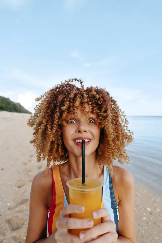 Young woman with curly hair drinks orange juice through a straw on a sandy beach under a bright sky, showing a playful and surprised expression.