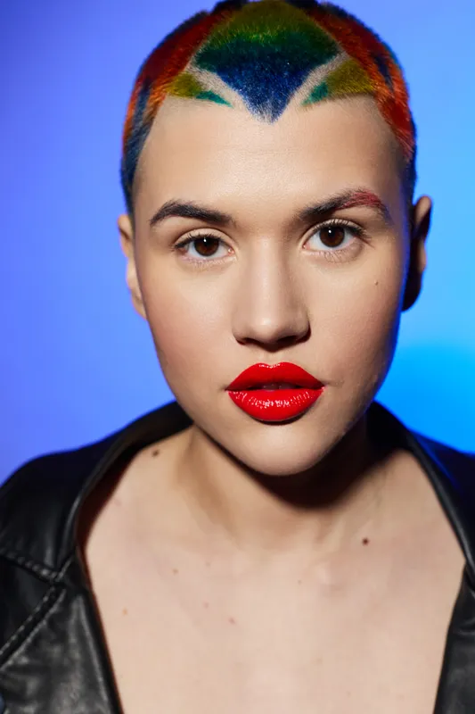 Bold woman with colorful rainbow hair and glossy red lips wears a black leather jacket against a vibrant blue background, portraying strength and uniqueness.