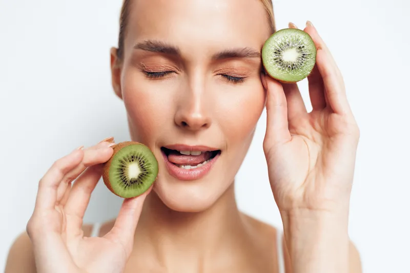 Young woman with clear skin holding two halves of kiwi fruit near her face with eyes closed enjoying fresh healthy lifestyle and natural beauty concept in bright studio light.