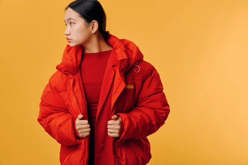 Young woman wearing red winter jacket and sweater looking sideways with serious expression on orange background. Studio portrait fashion model in warm clothes with tidy hairstyle and casual pose.