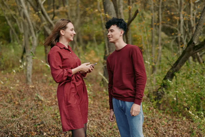 Young woman and teenage boy converse in a wooded clearing, smiling and holding a phone, natural gestures and genuine expressions conveying authenticity and credibility in a candid outdoor portrait.