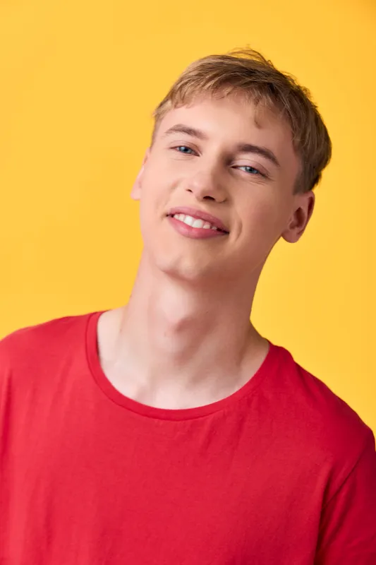 Young man in a red shirt smiles against a bright yellow background, casual lifestyle vibe with warm tones, relaxed pose and cheerful energy, suitable for fashion, marketing and lifestyle content.