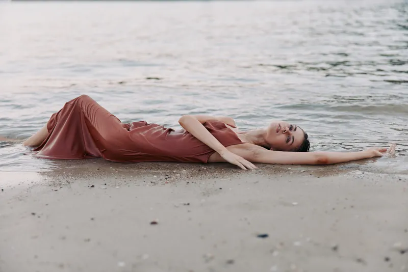 A serene figure lies on wet sand beside a calm sea, wearing a flowing red dress as gentle waves touch her silhouette, creating a dreamy, contemplative mood.