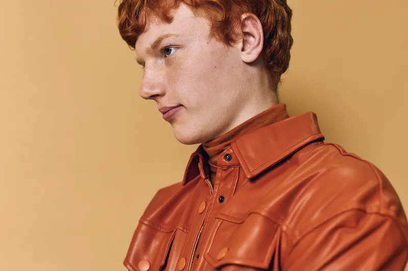 Portrait of young man with red hair wearing brown leather jacket against beige background. Profile view, casual fashion, natural expression, studio lighting, closeup headshot.