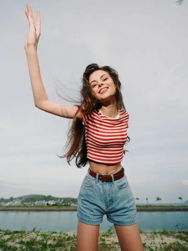Young woman outdoors wearing a red and white striped crop top and blue denim shorts raises her arms in joy, wind blowing her hair, bright sky and sunny summer mood.