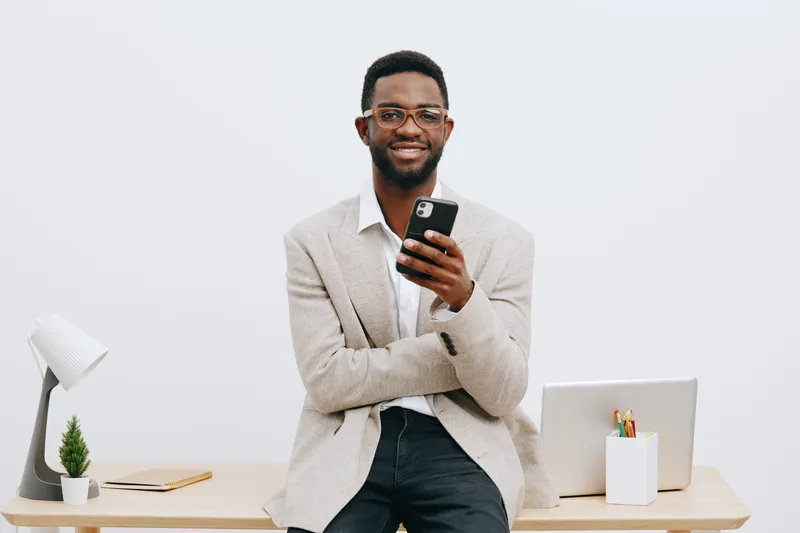 Confident smiling man using smartphone in modern office with minimalistic desk, laptop, plant, and lamp, dressed in casual blazer and jeans, positive work atmosphere. People lifestyle concept