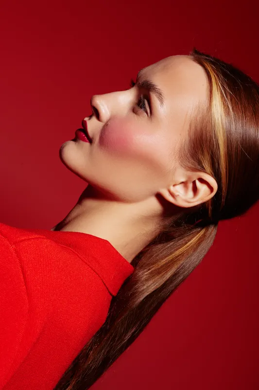 Profile of a young woman with smooth skin and red lipstick wearing a red sweater against a red background. Concept of beauty, fashion, and elegance in studio lighting.