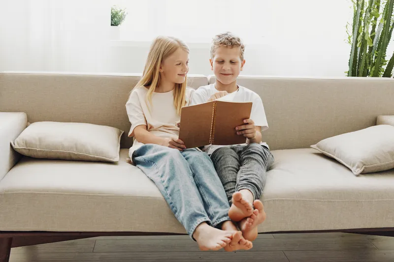 Two barefoot children sitting on beige sofa reading a book together in bright room with cozy cushions and plants, sharing learning moment.