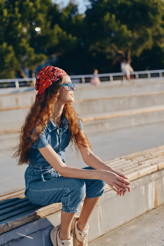 Lifestyle woman in modern boho-western denim street style wearing sunglasses and a red bandana, sitting outdoors in natural sunlight with film color effect.