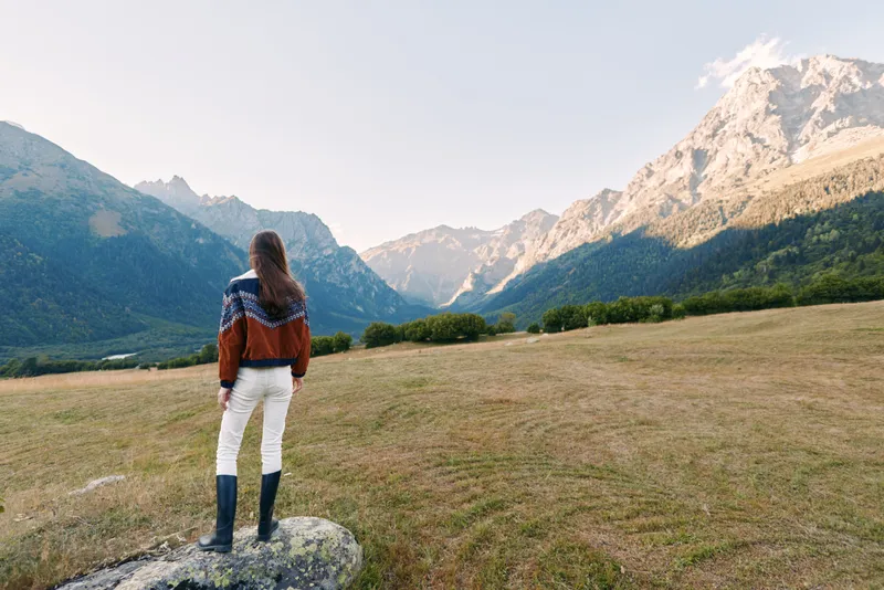 Woman in a patterned sweater stands on a rock in a wide meadow facing distant mountains, hiking across a peaceful landscape with boots, valley view and serene natural scenery.