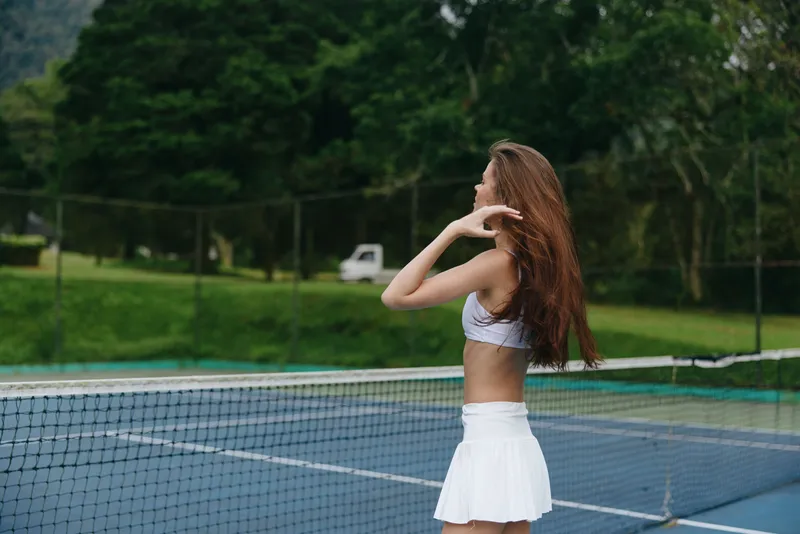 Young athletic woman in a sports outfit with long hair, enjoying the fresh air on a tennis court, showcasing confidence and vitality in a vibrant green setting