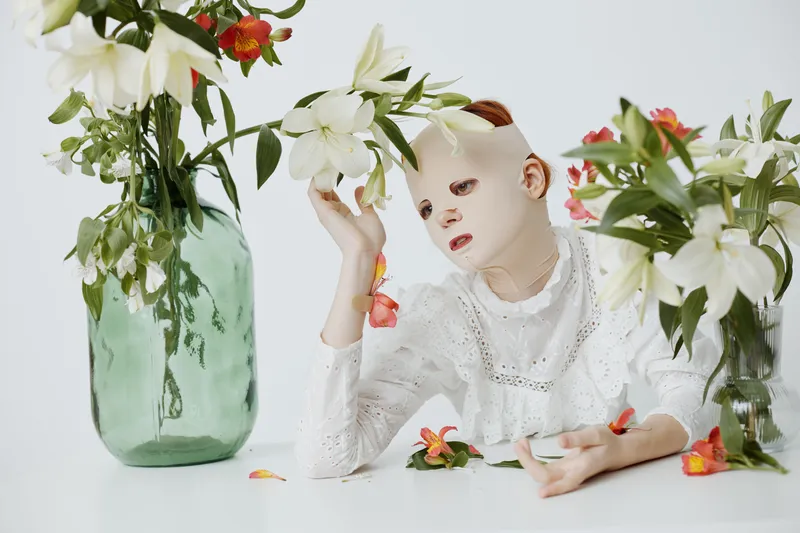 A serene model with a facial mask interacts delicately with white flowers in a minimalist setting, embodying themes of beauty, identity, and emotional depth
