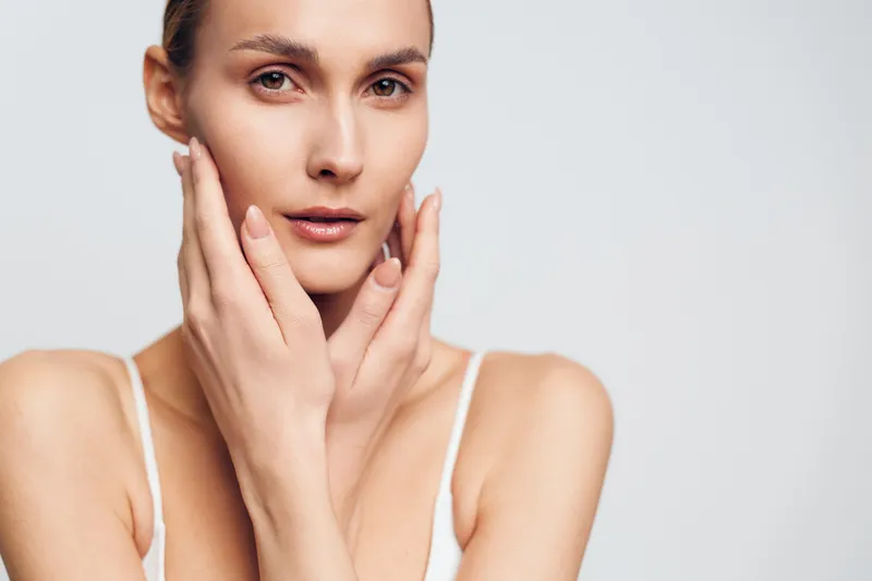 Young woman skincare beauty portrait, hands on face, calm expression, smooth skin, minimal spa vibe, soft studio light on neutral background, close up glamour, beauty routine relaxation
