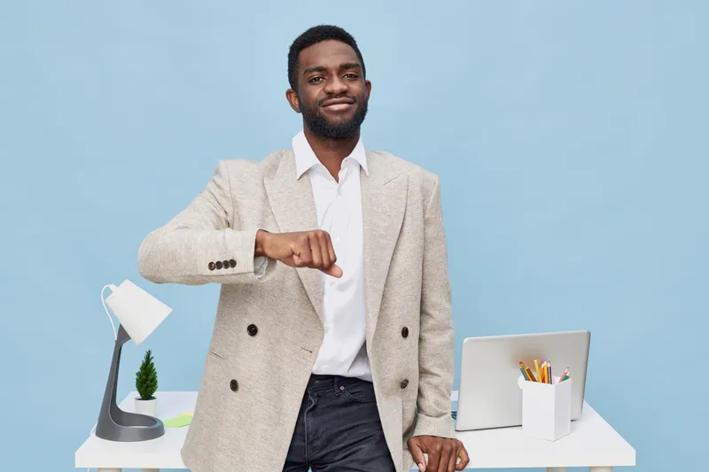 Portrait of a confident smiling man with dark skin and afro hairstyle, wearing a beige blazer and white shirt, standing in front of a modern desk with a laptop, desk lamp, and stationery, light blue b