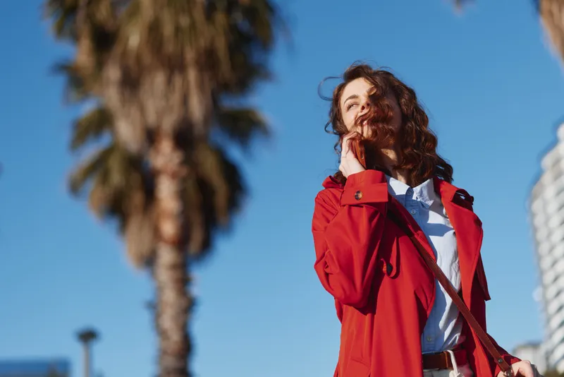 Young woman with curly hair wearing a red trench coat stands outdoors under a clear blue sky, smiling subtly as breeze moves her hair and fabric.