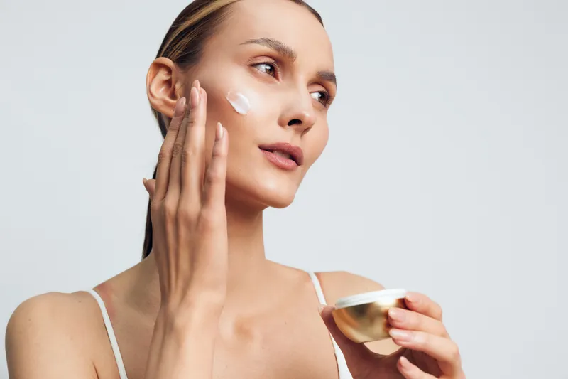 Young woman applies face cream on clean skin with fingers holds jar of moisturizer skincare beauty routine isolated on white background natural healthy complexion