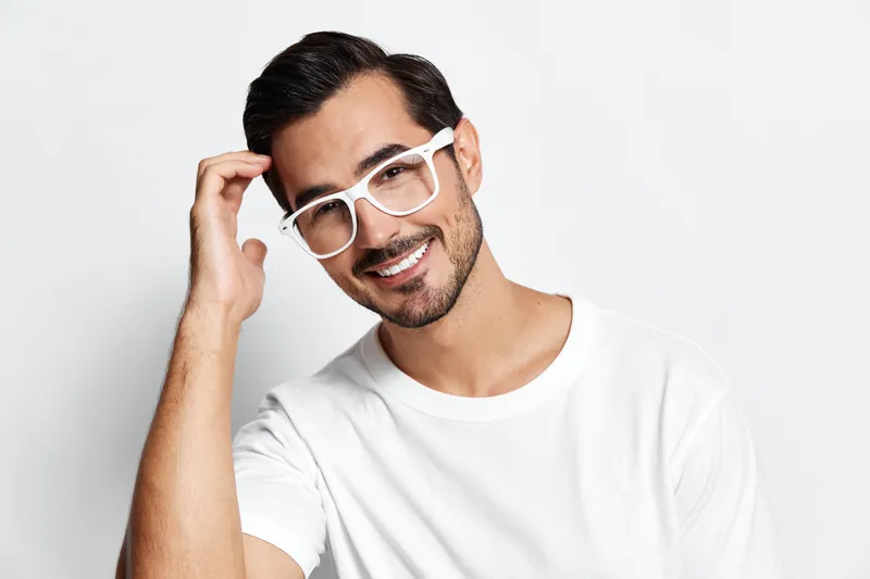 Smiling man with stylish glasses playfully poses while touching his hair, dressed in a casual white t-shirt, isolated on a light background. People lifestyle concept.
