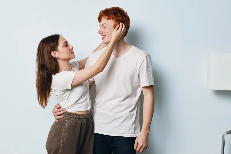 Couple, woman and man looking at each other, embracing and touching face in casual clothes at home. Relationship, affection and love with happy young people in simple interior.