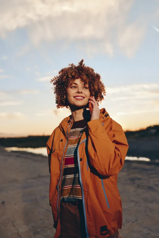 Woman with curly hair smiling outdoors at sunset, wearing a brown jacket and scarf, natural portrait, warm evening light, calm lifestyle moment, open landscape background