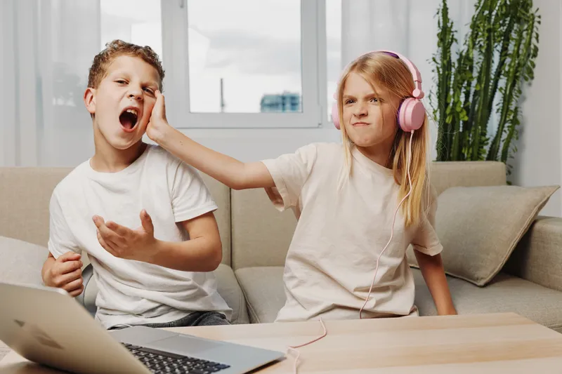 A playful girl with headphones pushes a boy’s face showing a funny annoyed expression sitting on a couch. The scene captures sibling interaction and childhood emotions indoors.