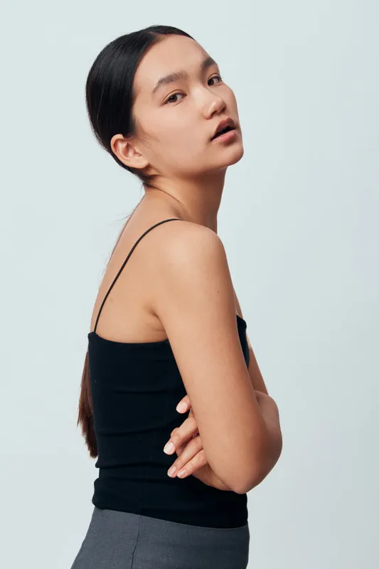 Young woman posing with folded arms wearing black tank top and gray pants against light background. Female model with long dark hair looking thoughtfully at camera in studio portrait.