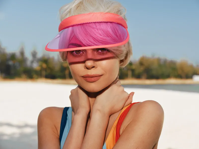 Portrait of a young model on a sunlit beach wearing a pink visor and colorful swimsuit. Soft light highlights skin, vibrant hair, and a calm, confident expression.