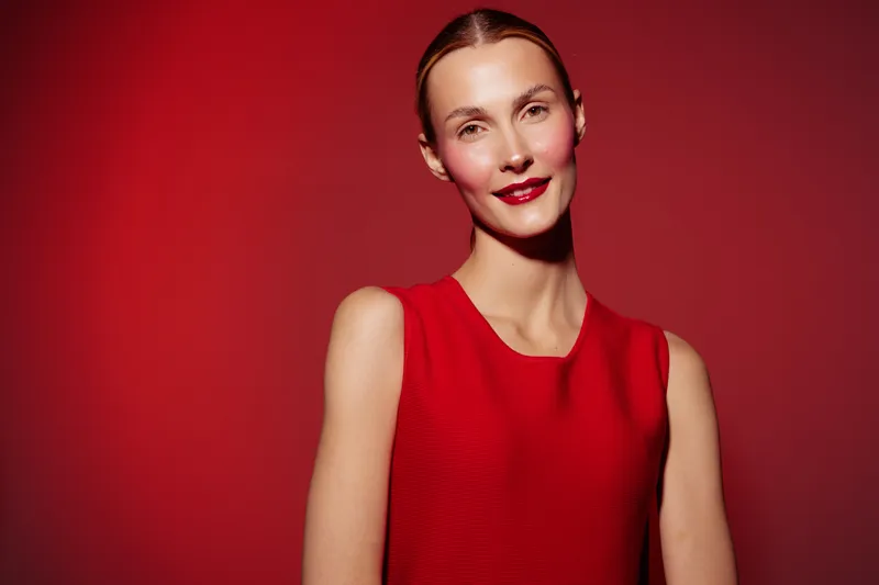 Smiling woman in red sleeveless dress poses against red background. Studio portrait with soft lighting highlights natural skin and confident expression. Concept for fashion and beauty.