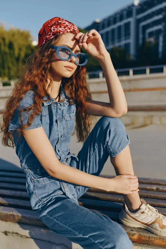 Lifestyle woman in modern boho-western denim street style with retro film color wearing a red bandana and blue sunglasses, seated casually outdoors during daytime.