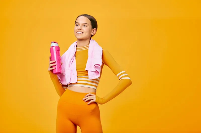 Fitness themed portrait of a woman in bright orange activewear, holding a pink water bottle with a towel draped over her shoulders, posed against a vivid orange backdrop.