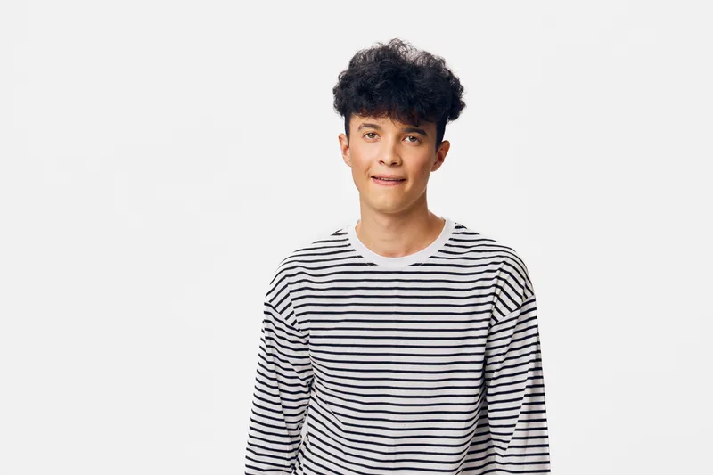young man in striped shirt with curly hair biting lip and looking at camera isolated on white background expressing thoughtful mood and casual style