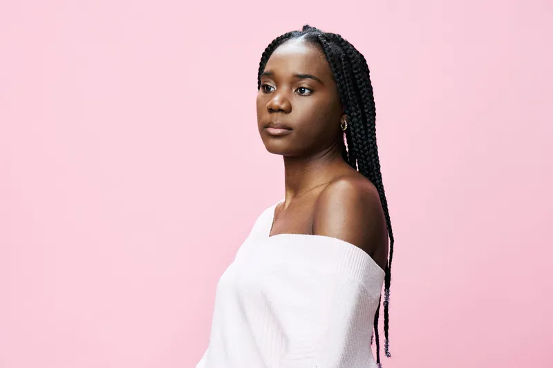 Young Black woman with braids, wearing a cozy sweater, poses thoughtfully against a vibrant pink background, radiating confidence and tranquility