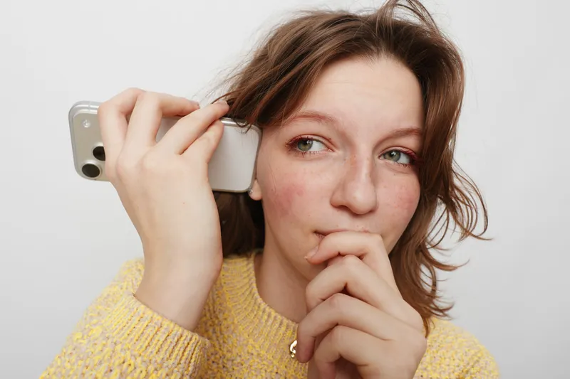Young woman with natural skin and freckles holds smartphone to ear, looking thoughtful. Casual style with yellow sweater on white background. Concept of communication and contemplation.