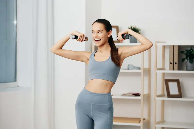 Woman fitness enthusiast in a bright home gym, wearing gray activewear, lifting dumbbells with a smile to show energy, balance, and confident movement during a workout.