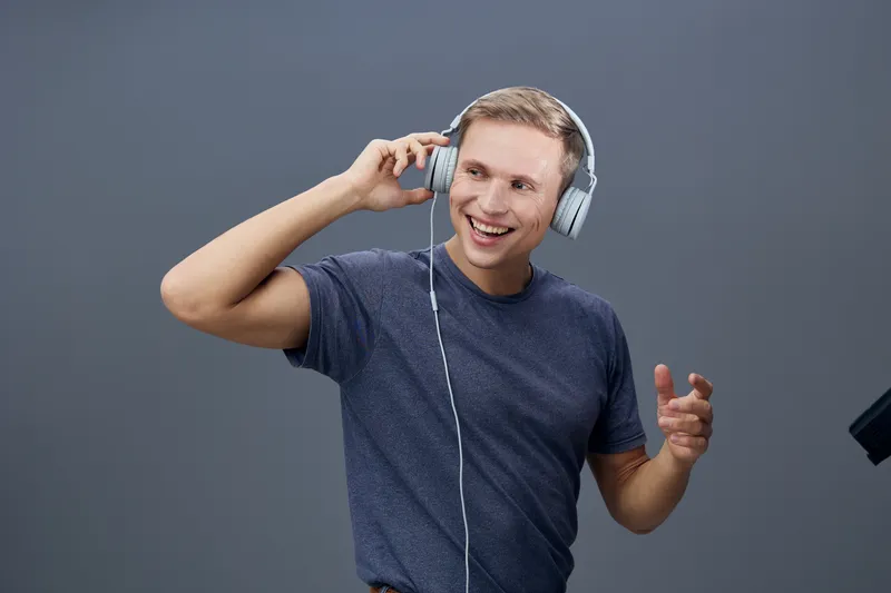 Happy young man listening to music with headphones smiling and gesturing against plain gray background. Casual blue t-shirt, cheerful expression, lifestyle and leisure concept.