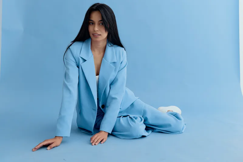 Woman in blue suit sitting on floor with calm expression and long black hair, wearing casual stylish outfit against a soft blue background.