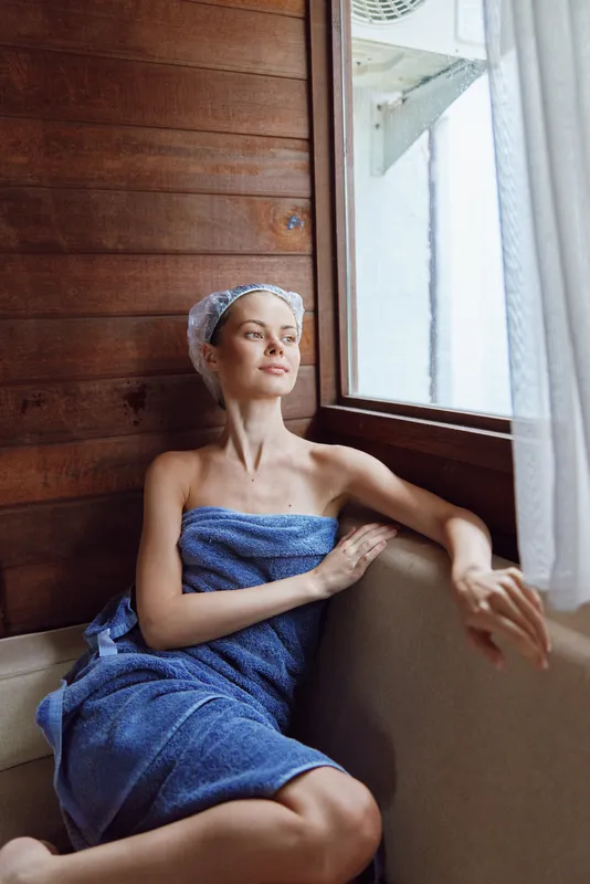 relaxed woman wrapped in blue towel sits by window in cozy wooden sauna, enjoying calm atmosphere and natural light on her peaceful face.