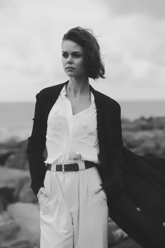 Fashionable woman stands on a windy coast wearing a white shirt, dark blazer and belted trousers, exuding timeless elegance and confident poise by the sea in monochrome