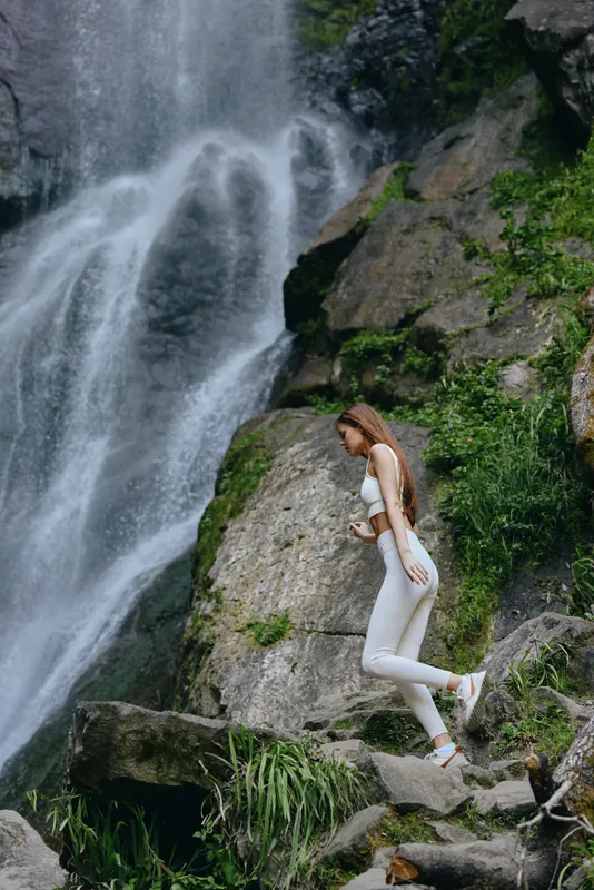 woman hiking near waterfall in white outfit exploring rocky terrain surrounded by greenery and flowing water
